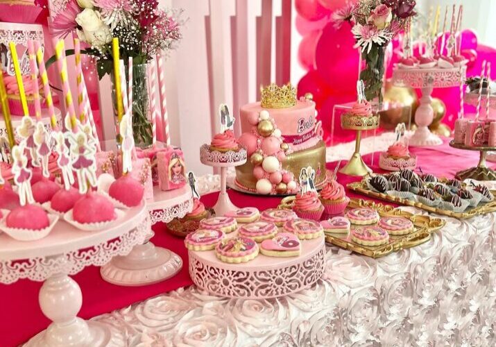 Barbie-themed dessert table with pink cakes, cupcakes, cookies, and character toppers