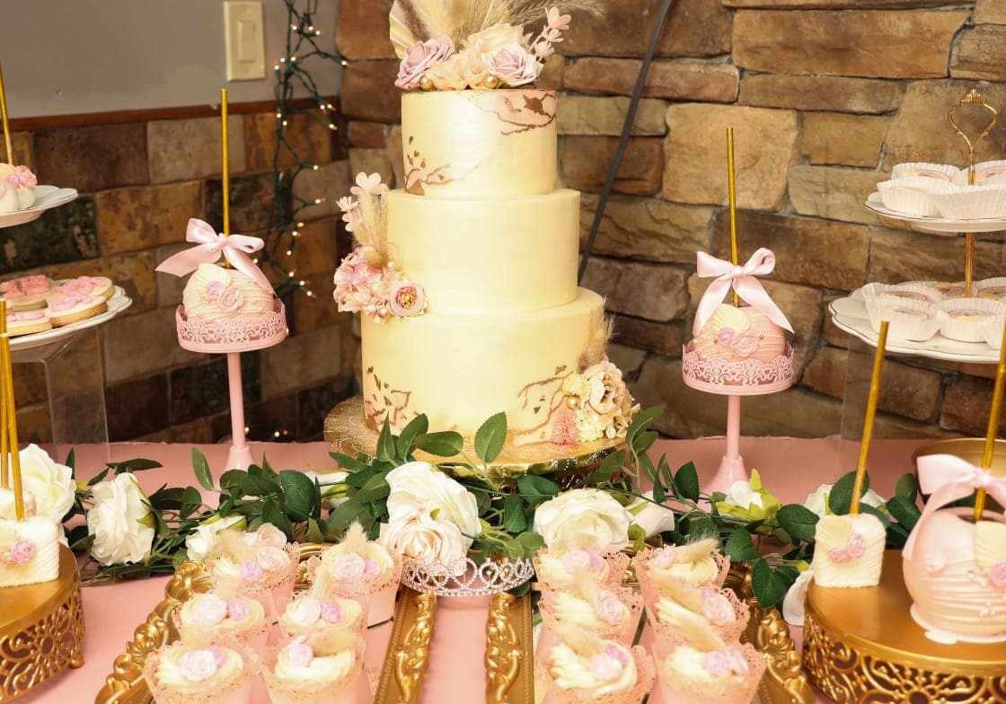 Rustic gold and blush three-tiered cake with floral decorations and coordinating desserts on a pink table