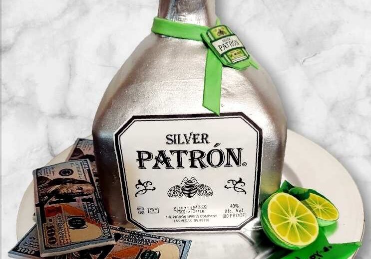 Silver Patrón bottle-shaped cake with fondant lime slices and edible dollar bills on a white plate